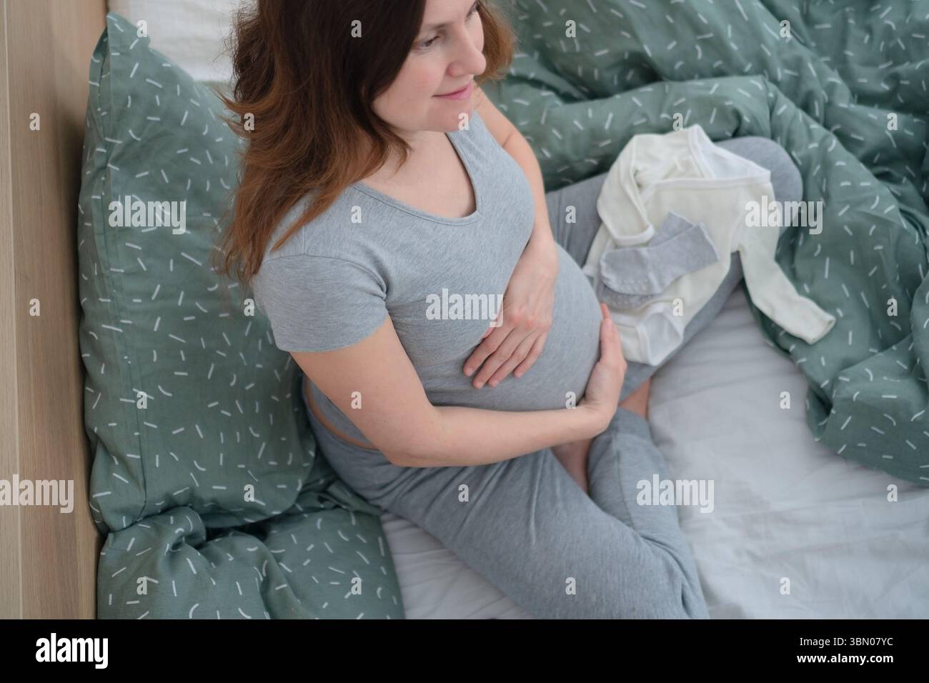 Una donna incinta incinta si siede a gambe incrociate, circondata da vestiti per bambini, toccando delicatamente la pancia. Un momento tranquillo di amore e anticipazione. Foto Stock