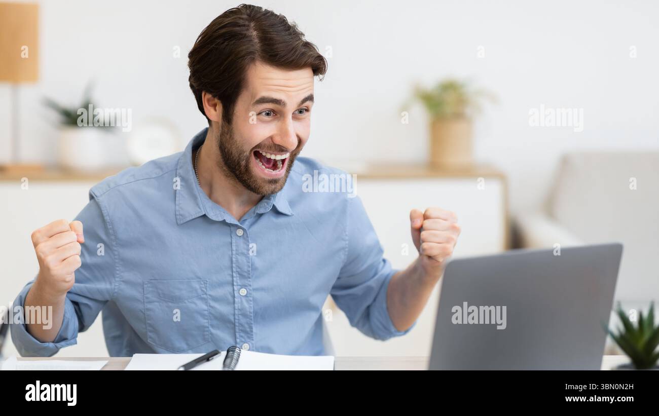 Eccitato giovane uomo d'affari al computer portatile scuotendo i Fists in ufficio Foto Stock