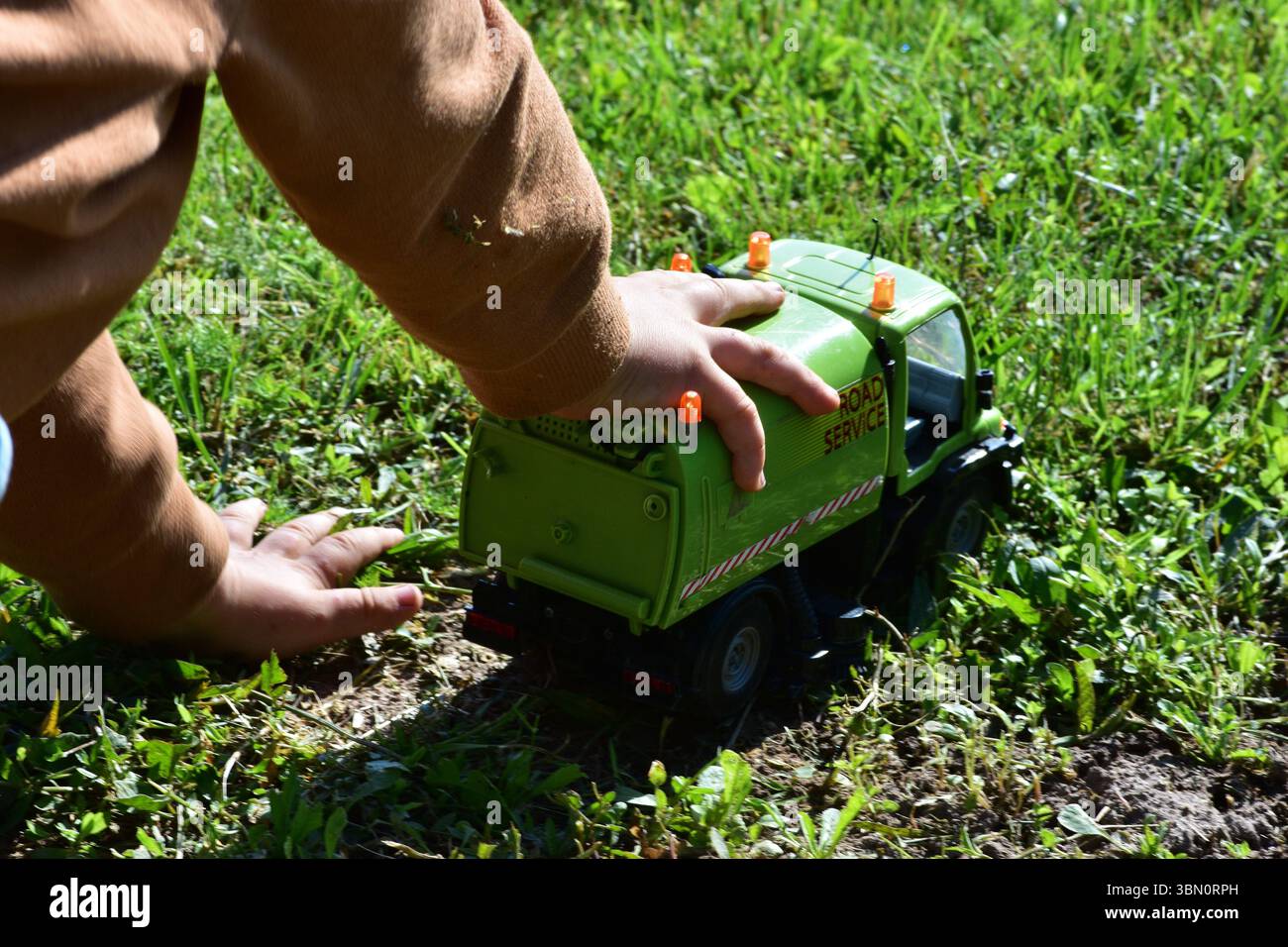 Il bambino gioca con un camion verde giocattolo su un prato erboso. Le mani del bambino spingono delicatamente il veicolo nelle giornate di sole. Tempo di gioco all'aperto pieno di Foto Stock