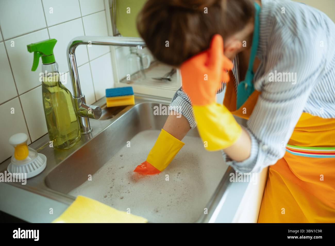 Donna in grembiule e guanti che pulisce un lavandino pieno di acqua saponata, probabilmente affrontando un frustrante problema di intasamento del lavandino della cucina. Foto Stock