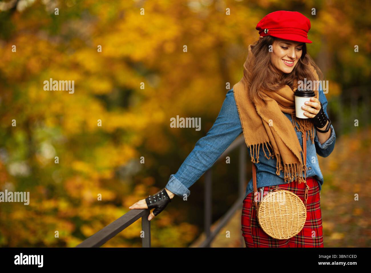 Ciao autunno. donna moderna sorridente in camicia jeans e cappello rosso con sciarpa, guanti, borsa e caffè nel parco cittadino. Foto Stock