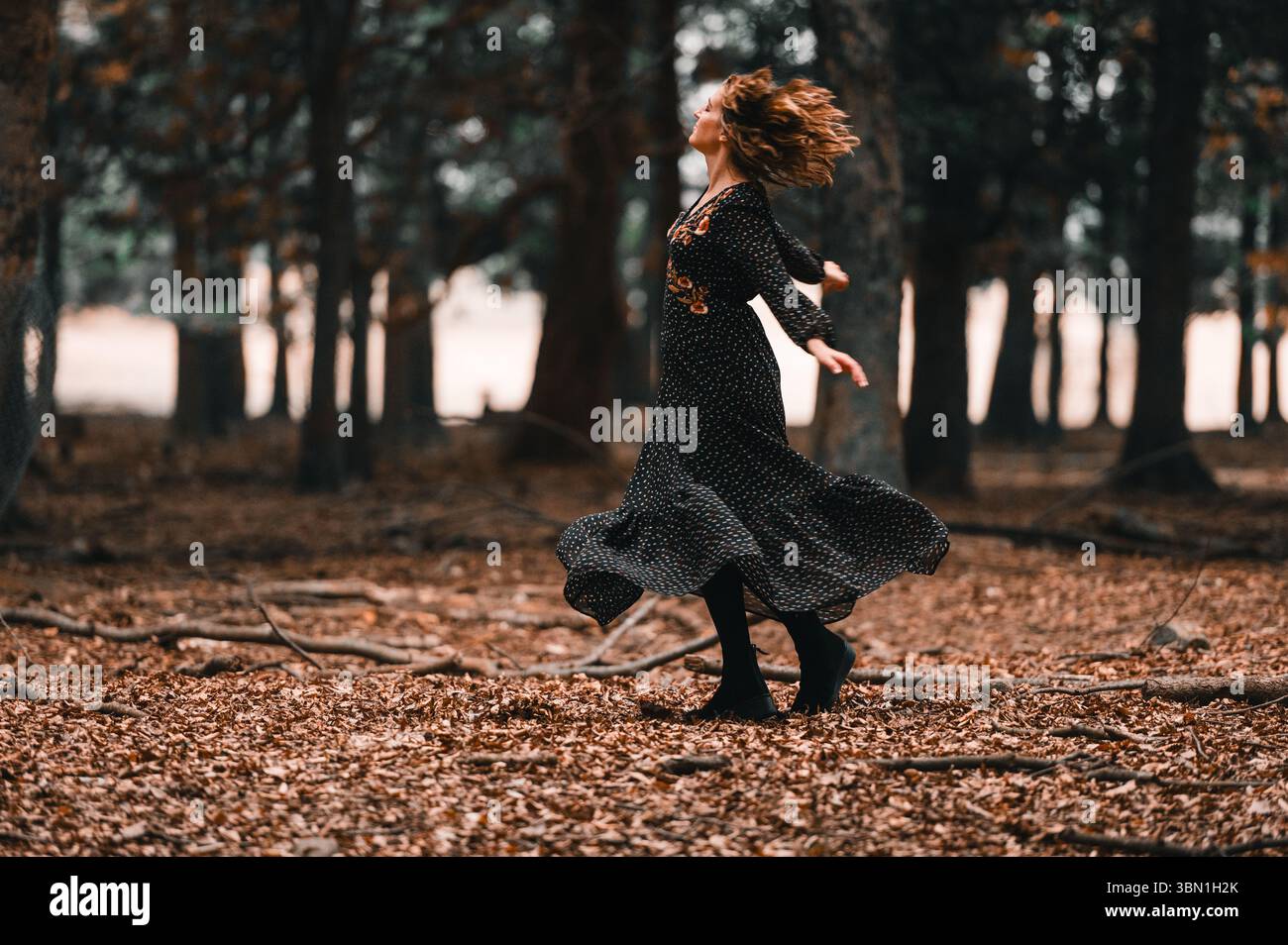 Donna che danzava nella foresta con foglie autunnali e un lungo abito floreale Foto Stock