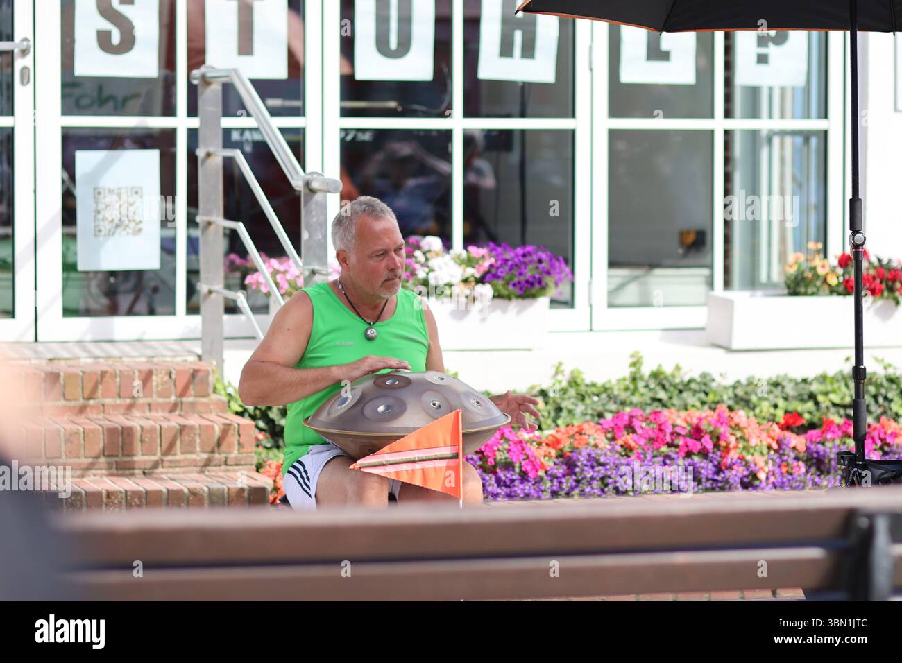 Amrum, Germania - 5 agosto 2024: Un musicista suona sapientemente un handpan in un vivace giardino urbano, circondato da una varietà di fiori in fiore. Sunligh Foto Stock