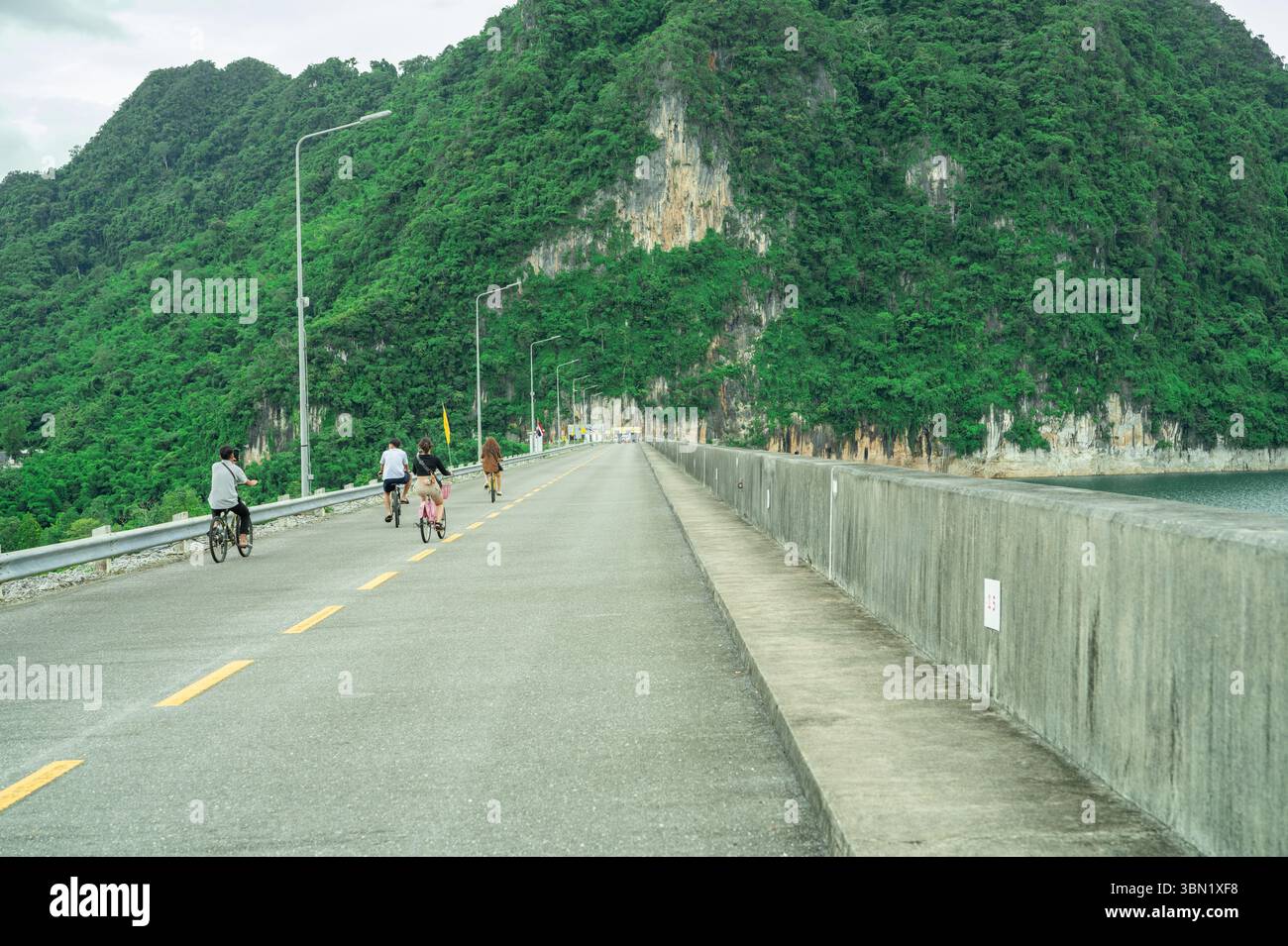 La famiglia pedala insieme lungo una strada di cemento su una diga panoramica circondata da scogliere calcaree durante il pomeriggio, promuovendo un turismo ecologico Foto Stock