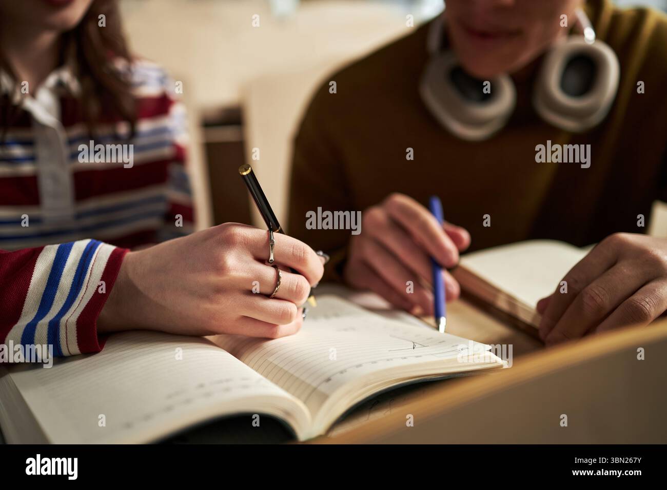 Giovane donna caucasica e ragazzo caucasico che studiano insieme all'università, scrivono appunti in quaderni, si concentrano sul lavoro accademico, sul primo piano delle mani e sui libri sulla scrivania Foto Stock