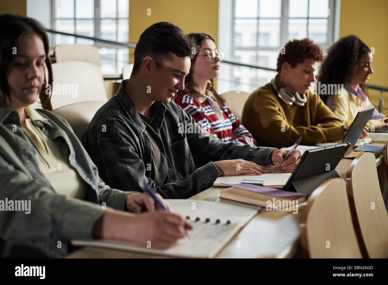 Gruppo di studenti universitari diversi seduti in una sala conferenze prendendo appunti e utilizzando tablet digitali, giovani tra cui individui caucasici e neri che si concentrano sul lavoro accademico durante le lezioni Foto Stock