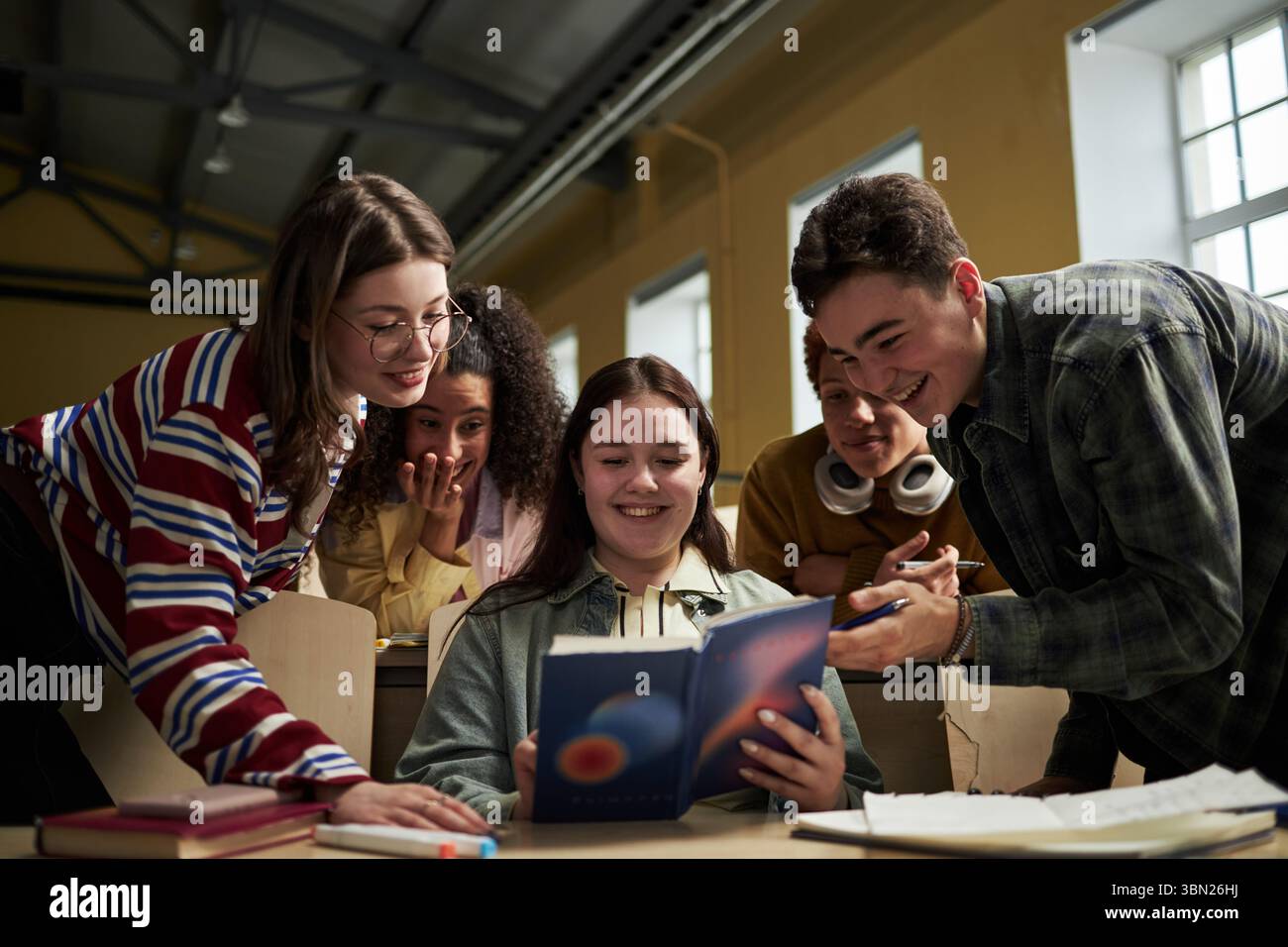 Gruppo di studenti universitari multietnici che si riuniscono intorno alla scrivania sorridendo e discutendo insieme libri di testo, giovani adulti che collaborano a progetti accademici in aula Foto Stock