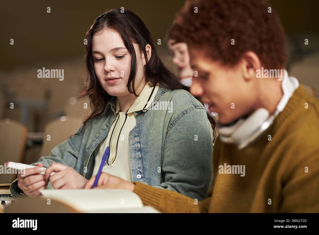 Giovane donna caucasica e giovane nero che studiano insieme in classe universitaria, scrivono appunti e discutono di compiti, entrambi focalizzati sul lavoro accademico, offuscano lo studente in background Foto Stock