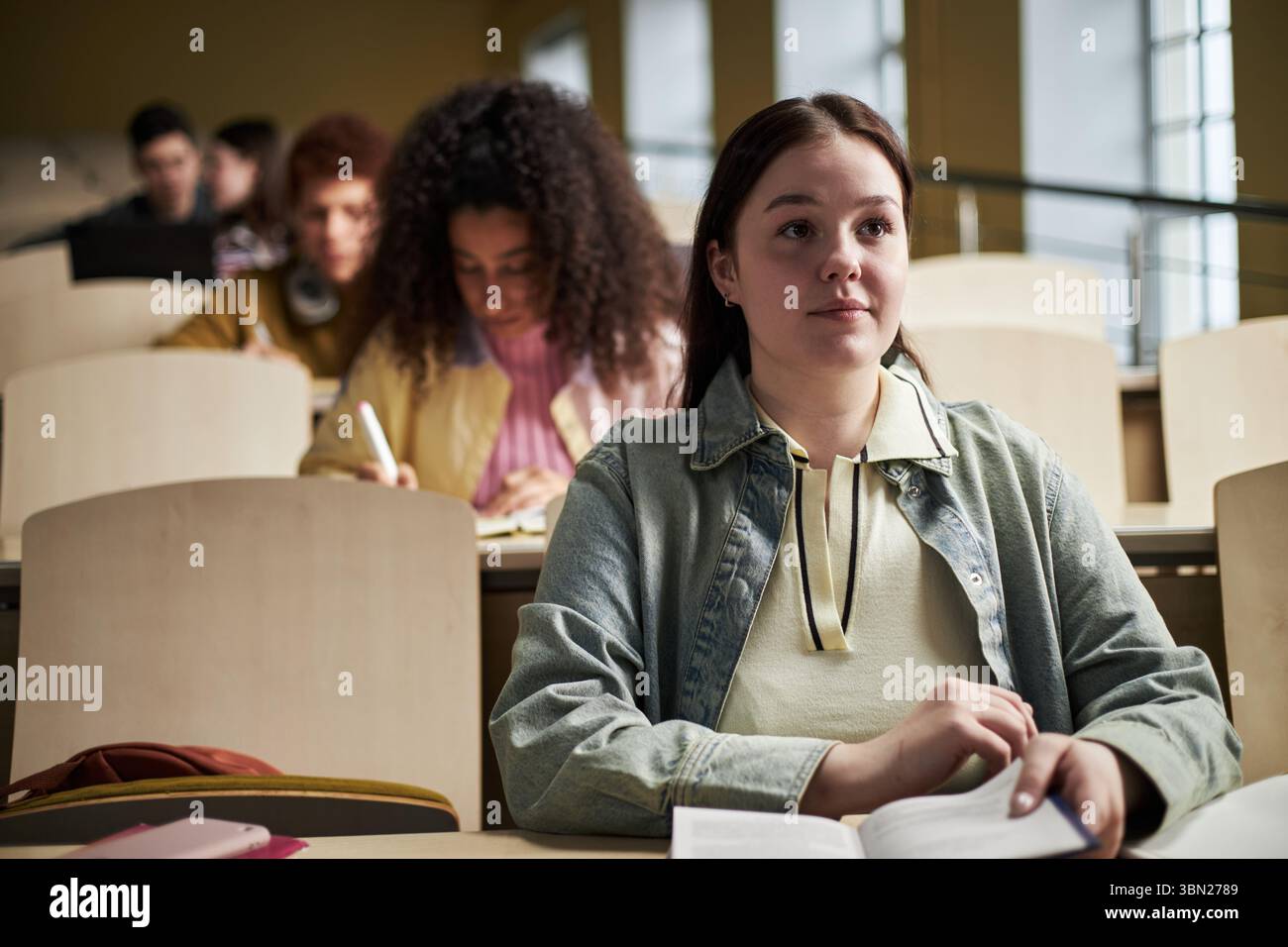 Felice giovane donna caucasica seduta in una sala conferenze universitaria che tiene un libro aperto, guardando avanti, gruppo multietnico di giovani studenti che studiano e scrivono in background Foto Stock