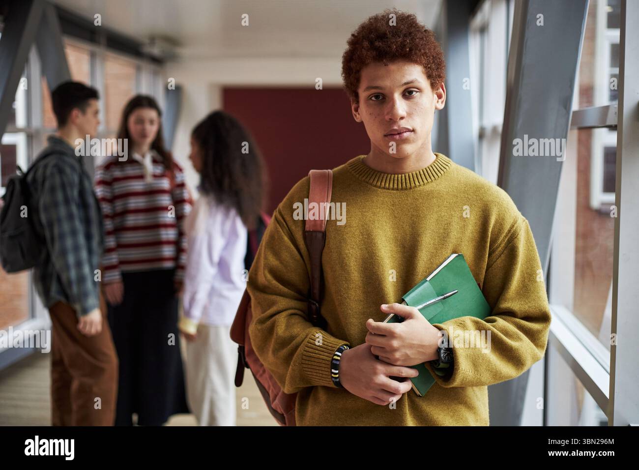 Ritratto di un giovane uomo di colore in piedi nel corridoio dell'università con notebook e zaino, guardando la fotocamera mentre un gruppo di studenti multietnici parla in sottofondo Foto Stock