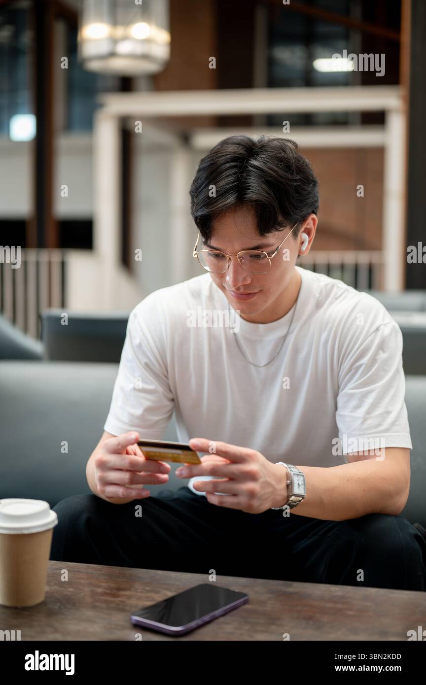 L'uomo asiatico che indossa occhiali in maglietta bianca sta guardando una carta di credito gialla che tiene con entrambe le mani. Pagamento con carta, pubblicità bancaria. Foto Stock