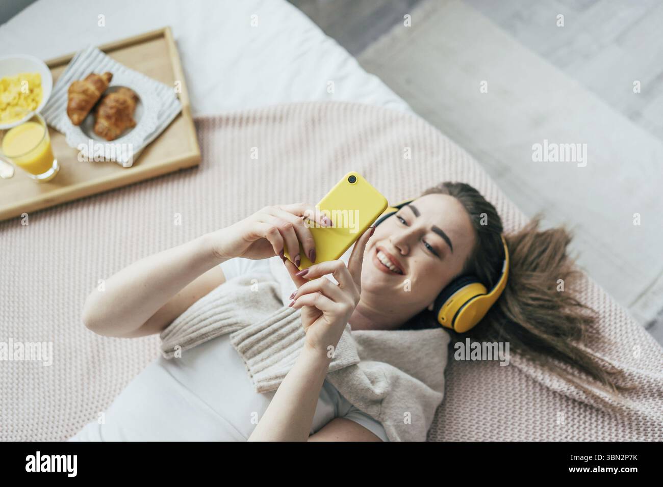 Giovane e attraente bruna sorridente con le cuffie gialle è sdraiata sul letto, tenendo in mano un telefono e ascoltando musica Foto Stock