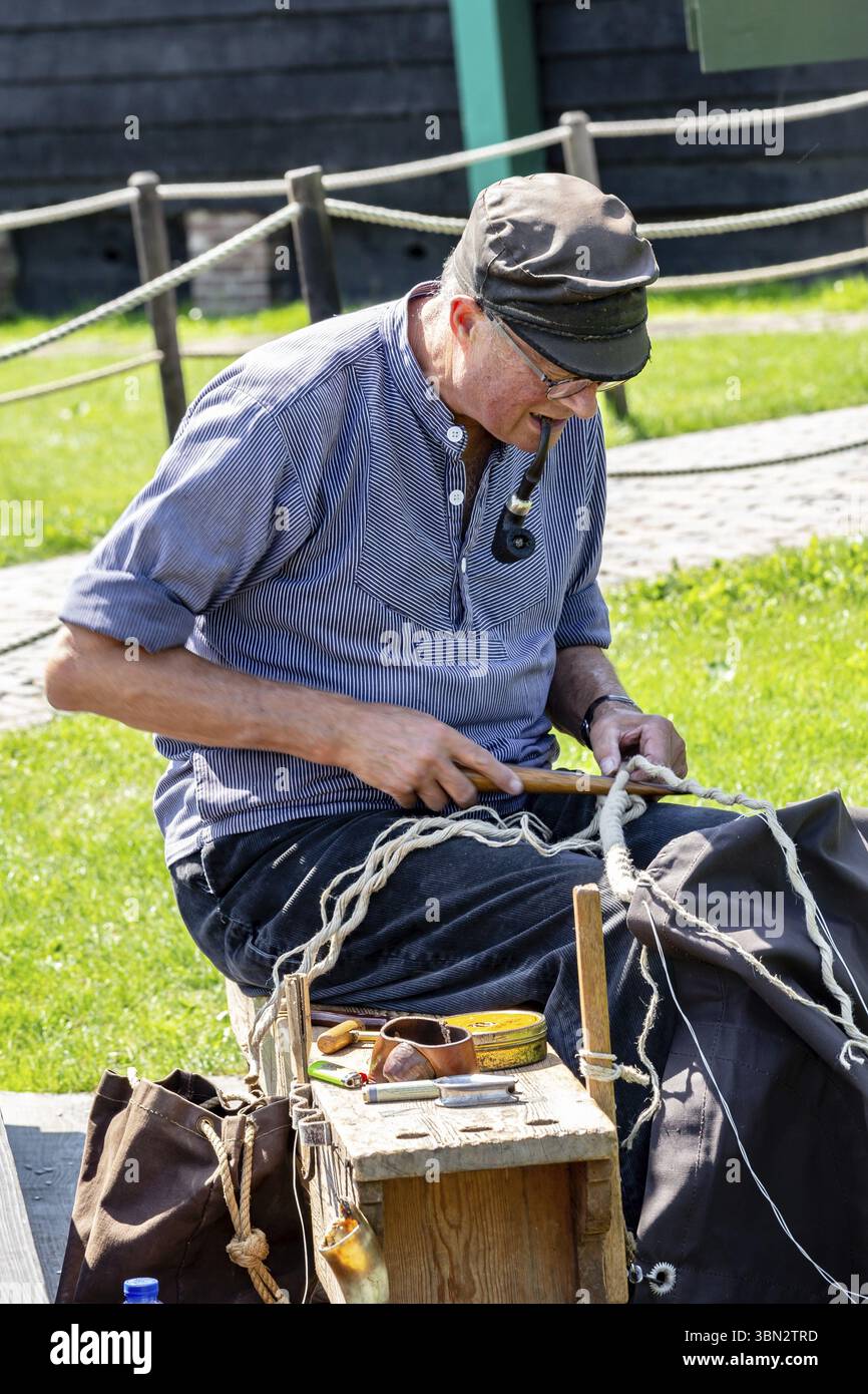 Zaanstad, Paesi Bassi - 18 luglio 2021: Miller con tubi e abbigliamento tradizionale che lavorano su corda di fronte al mulino a vento in Zaanse Schans Village in Foto Stock