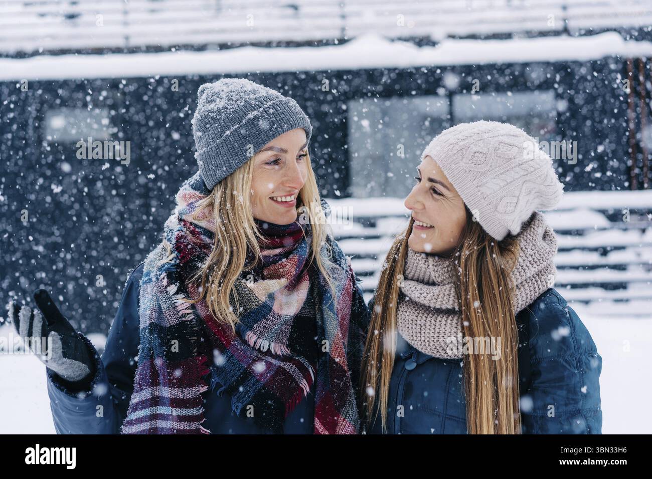 Ritratto di due amiche donne in cappelli e giacche in piedi sotto la neve e che si guardano sorridendo Foto Stock