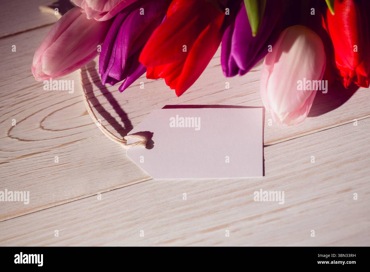 Il gruppo di tulipani proietta ombre morbide sul piano portapaziente in legno dal design piatto, con etichetta regalo vuota Foto Stock