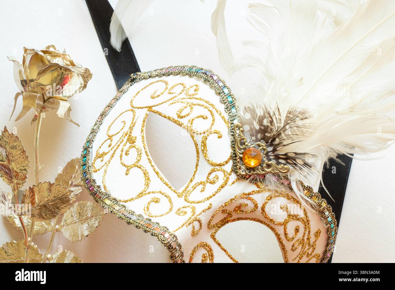 Maschera bianca di carnevale con piume bianche e una rosa dorata accanto ad essa su sfondo bianco, accessorio decorativo per una maschera Foto Stock