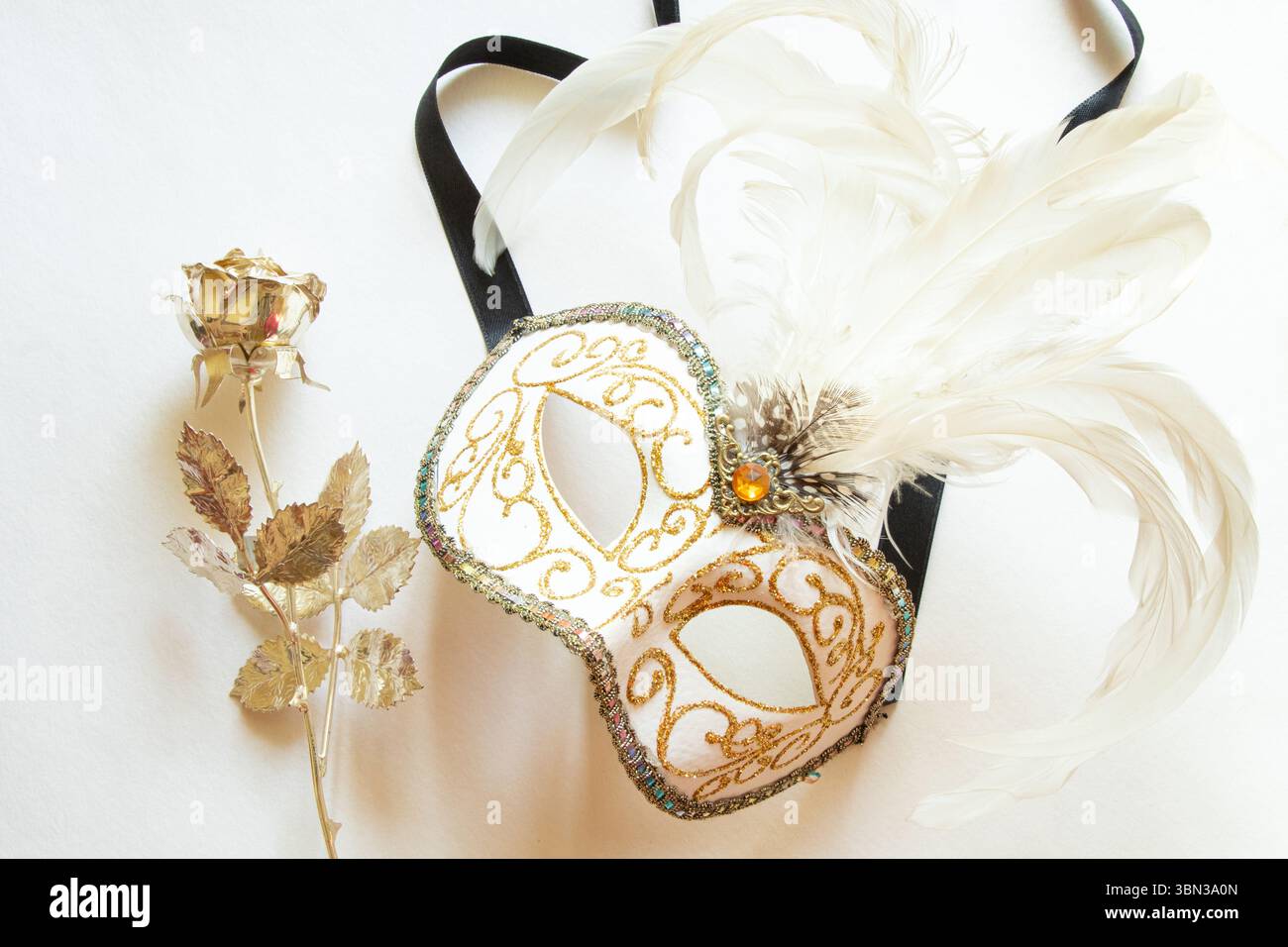 Maschera bianca di carnevale con piume bianche e una rosa dorata accanto ad essa su sfondo bianco, accessorio decorativo per una maschera Foto Stock