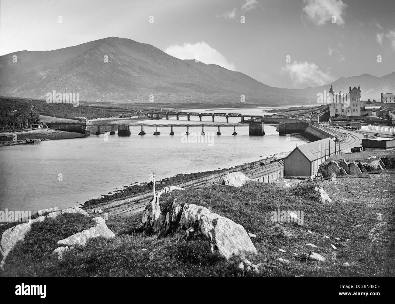 Una fotografia del tardo XIX secolo della stazione ferroviaria e dei ponti stradali e ferroviari che attraversano il fiume Ferta a Cahirciveen sulla penisola di Iveragh, nella contea di Kerry, in Irlanda. La stazione Royal Irish Constabulary (RIC), progettata in modo flambolescente 1870, fu costruita nel caratteristico stile "Schloss" favorito dal suo architetto, Enoch Trevor Owen, e ora un centro storico. Foto Stock