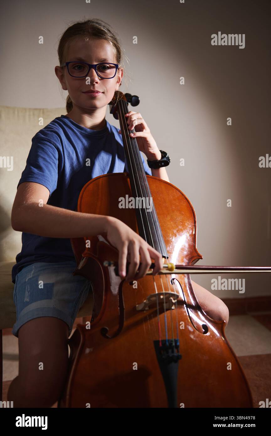 Un giovane violoncellista che indossa un abbigliamento casual pratica al chiuso sotto un'illuminazione mirata, mettendo in evidenza la loro abilità e dedizione alla musica in un ambiente calmo Foto Stock