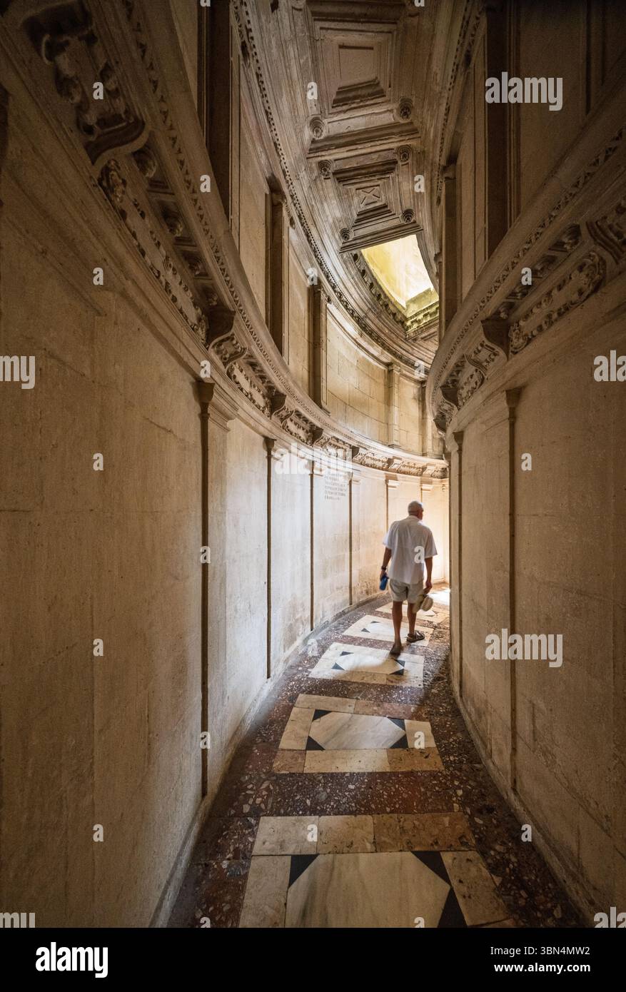Un visitatore cammina attraverso un corridoio curvo all'interno della Cattedrale di Siviglia, mostrando pareti ornate e un pavimento piastrellato a motivi geometrici che conduce alla sala. Foto Stock