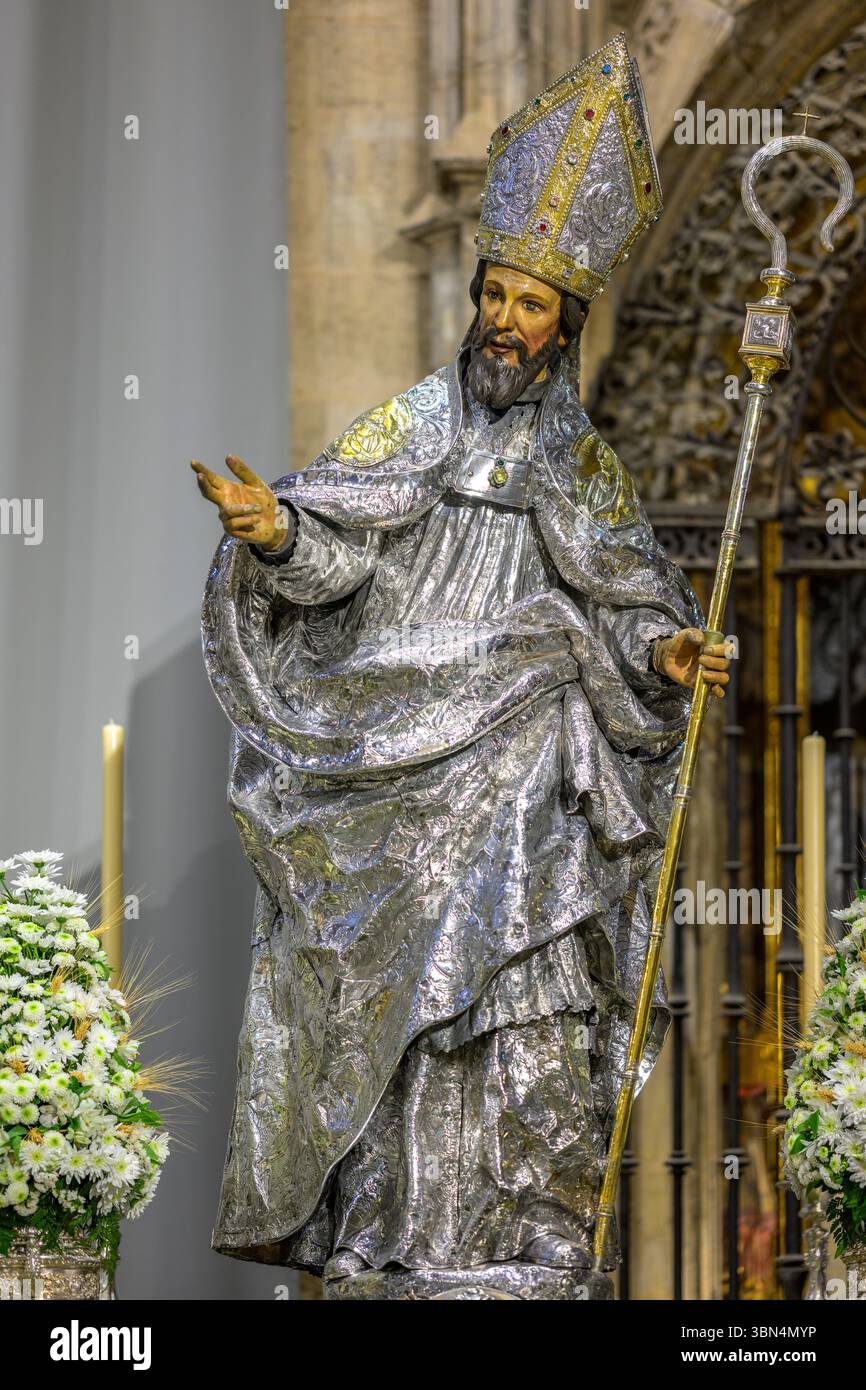 Una scultura policroma di San Leandro adornata con paramenti episcopali si erge con un libro e un crozier, circondato da fiori e candele. Foto Stock