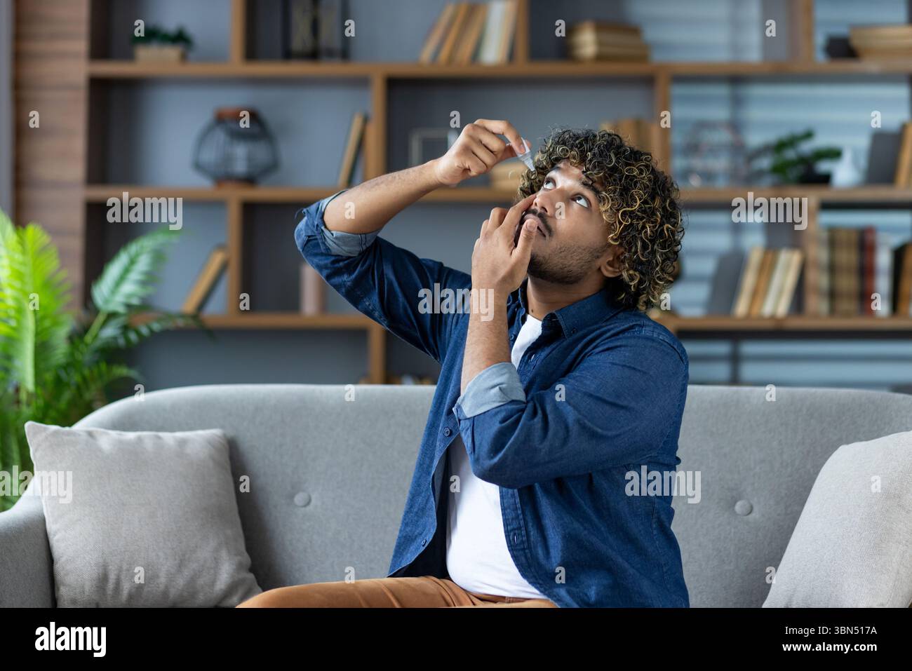Un uomo con i capelli ricci applica gocce d'occhio, seduto su un divano, con una libreria sullo sfondo. Sembra che stia vivendo disagio agli occhi. Foto Stock