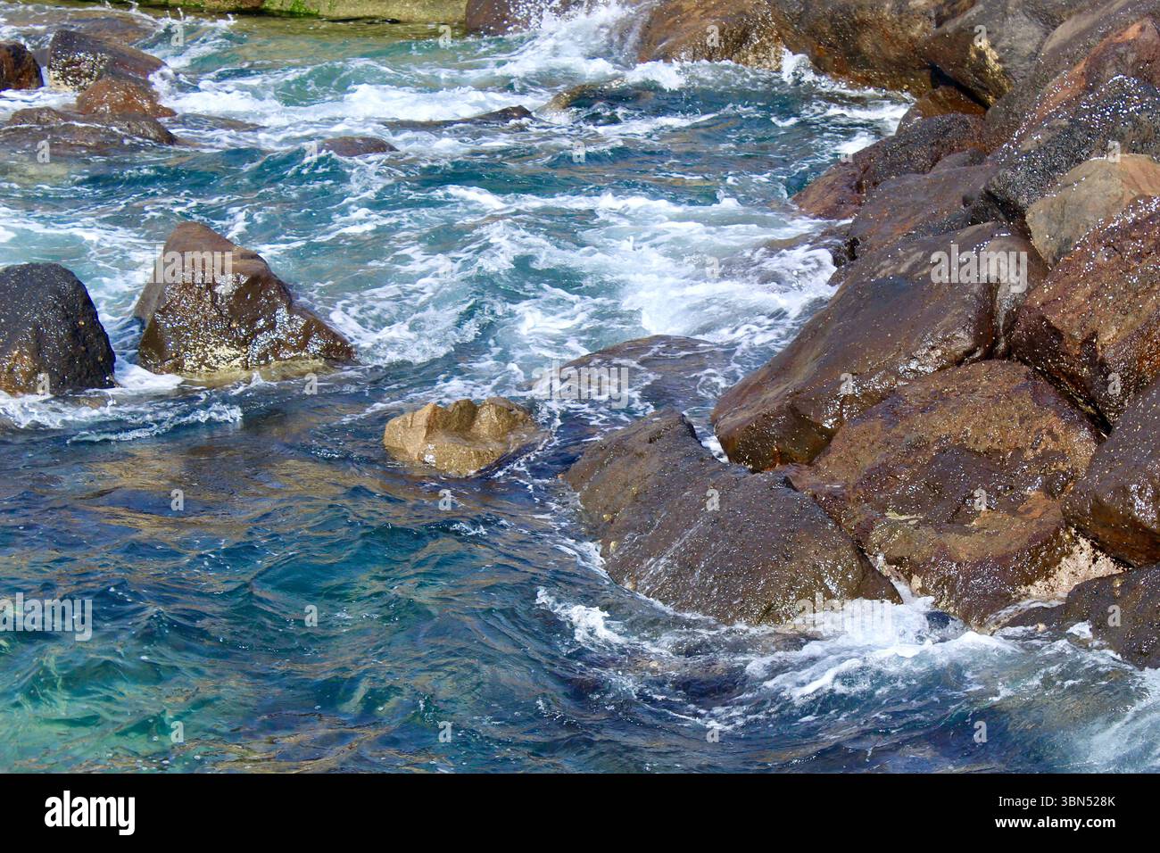 Vista ravvicinata delle onde oceaniche che si infrangono contro le aspre rocce costiere. Il turchese turchese e l'acqua di mare blu intenso creano uno spettacolare spruzzo bianco Foto Stock