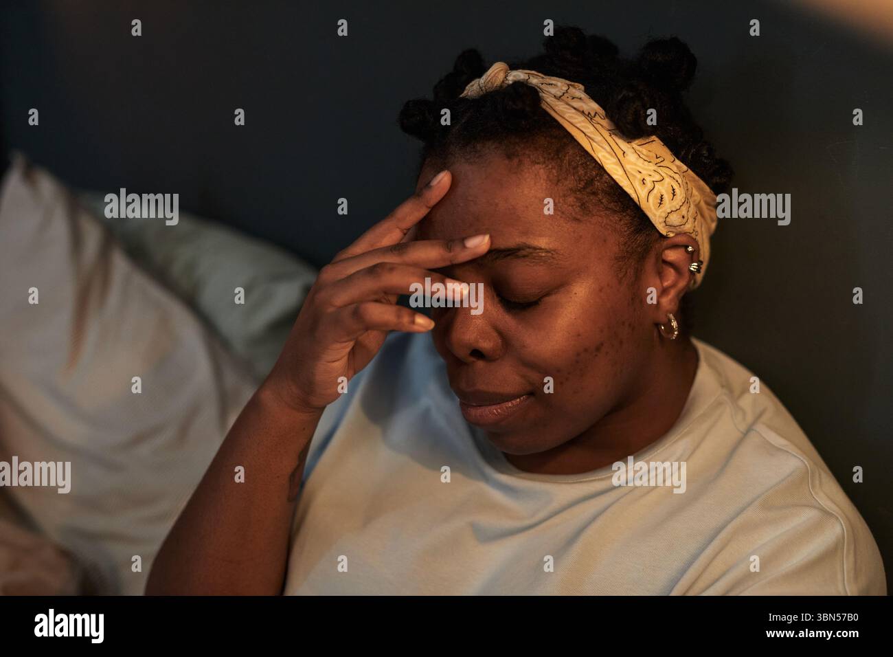 Donna di colore giovane adulto seduto sul letto toccando la fronte a occhi chiusi che mostrano stress o frustrazione durante situazioni di conflitto, testa leggermente inclinata, con archetto Foto Stock