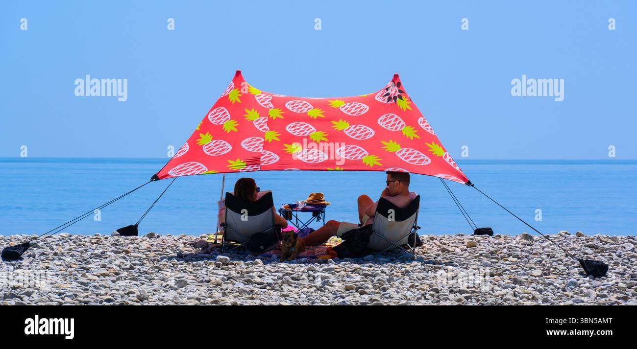 Lyme Regis, Dorset, Regno Unito. 30 giugno 2025. Meteo nel Regno Unito. La gente si riparava sotto ombrelloni dai colori vivaci mentre il caldo sole pomeridiano tramontava nella località balneare di Lyme Regis il giorno più caldo dell'anno finora. Le temperature sono destinate a salire bene a metà degli anni '30, mentre l'ondata di calore di giugno persiste. Crediti: Celia McMahon/Alamy Live News Foto Stock
