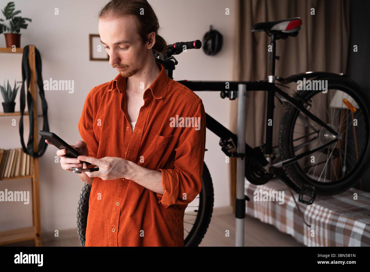 Uomo che ripara la bicicletta in ambienti interni, controlla lo smartphone per istruzioni e suggerimenti per la risoluzione dei problemi relativi alla manutenzione e all'autoriparazione della bicicletta. Concetto di bici da casa Foto Stock