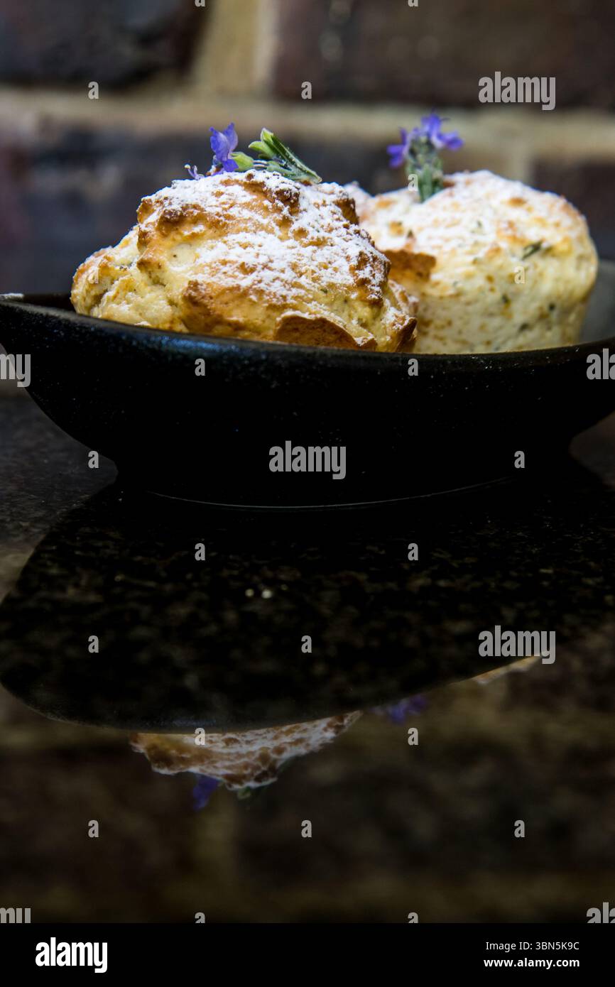 Muffin aromatizzati al rosmarino, decorati con una spolveratura di zucchero a velo e fiori di rosmarino viola, che si riflettono sul piano del tavolo in pietra scura, lucido e liscio, Foto Stock
