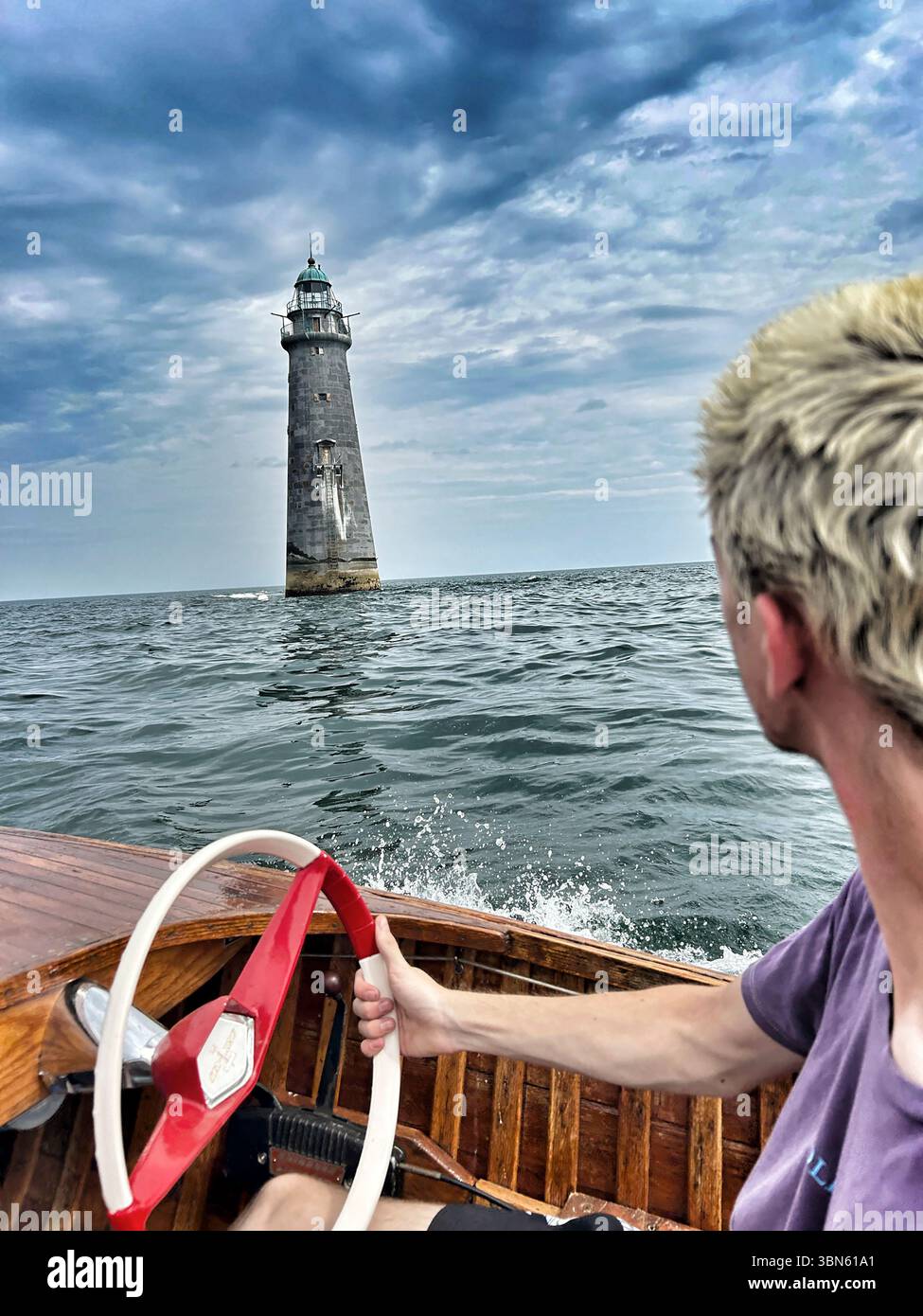 Giovane adulto che guida una barca a motore d'epoca vicino al faro Foto Stock