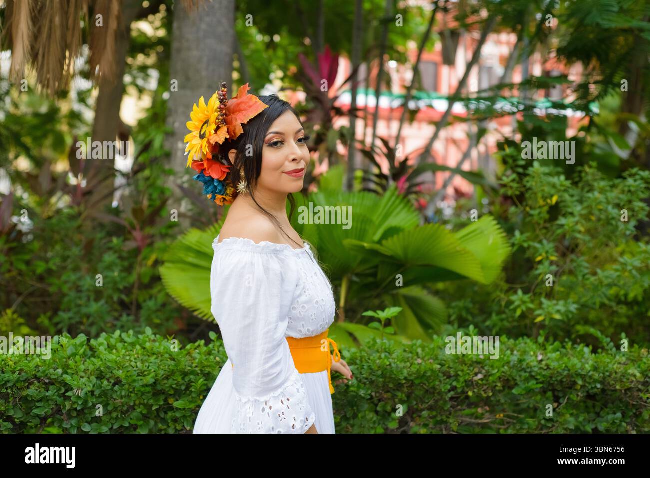Una donna con un abito bianco a spalla, decorato da un vivace copricapo floreale autunnale, si erge graziosamente in mezzo a una vegetazione lussureggiante. Foto Stock