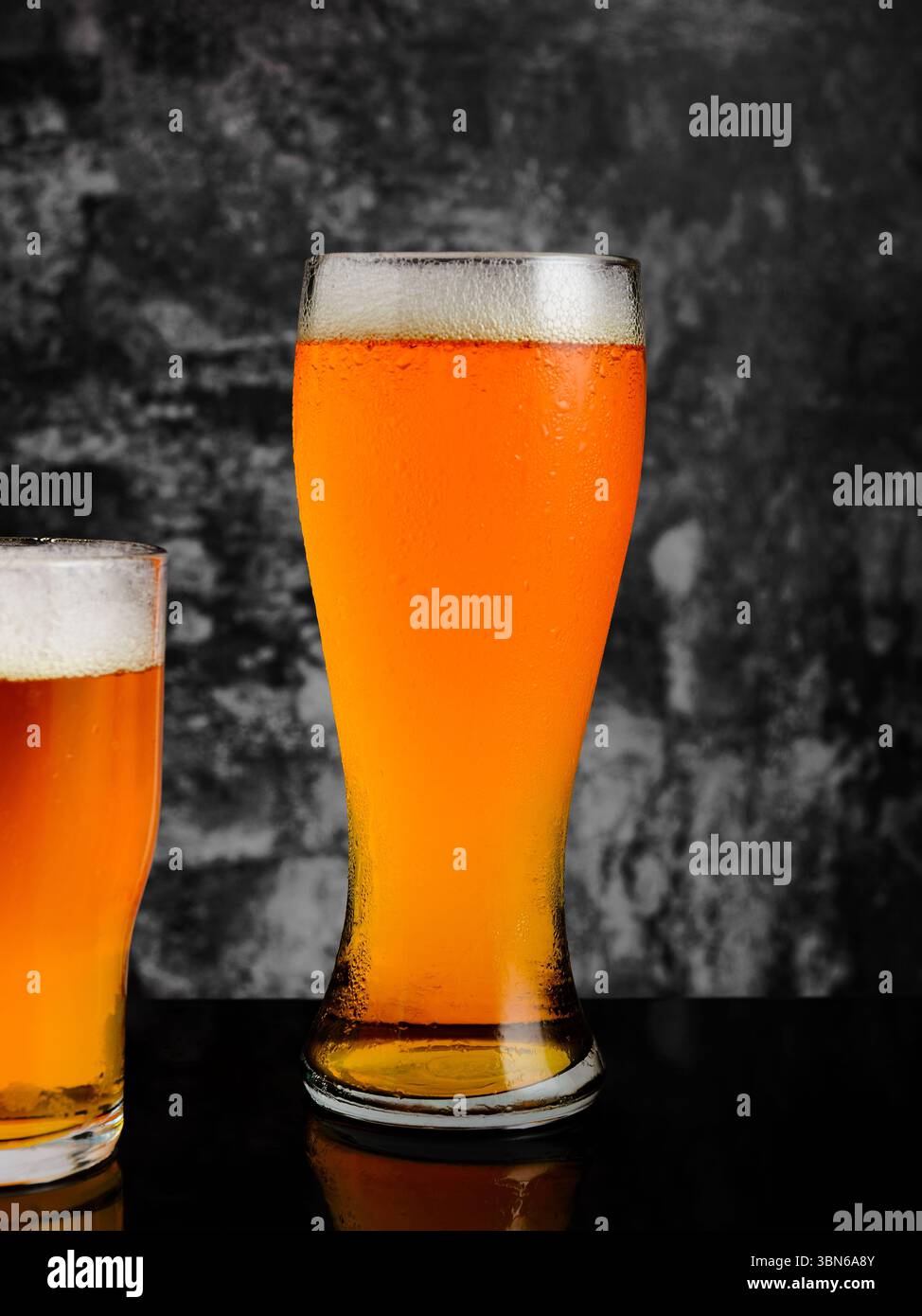 Due bicchieri di birra gialla, uno alto, uno corto, poggiano su uno sfondo scuro e strutturato. Foto Stock