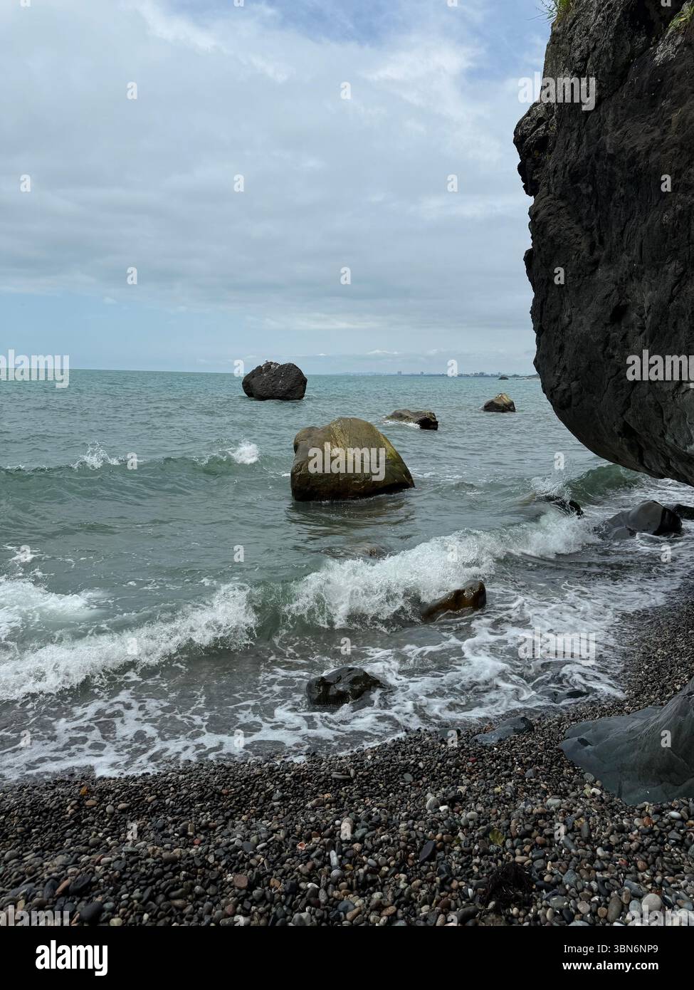 L'immagine mostra una spiaggia di ciottoli con piccole onde che colpiscono la riva. Nell'acqua sono visibili diverse grandi rocce, e un'alta scogliera è sul lato destro Foto Stock