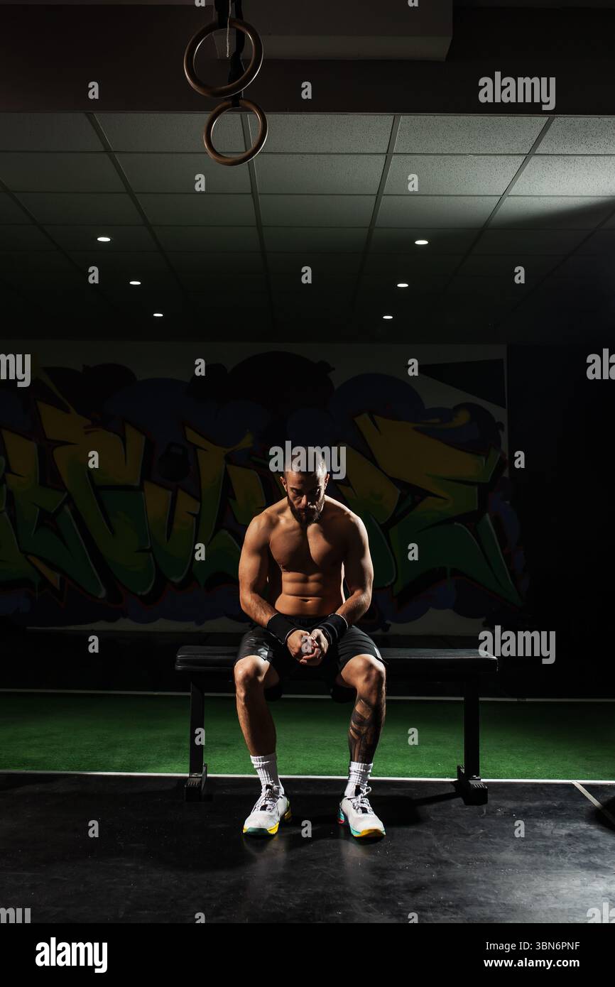 Atleta maschile tatuato e concentrato siede su una panchina in una palestra poco illuminata, fermandosi tra un allenamento e l'altro sotto una spettacolare illuminazione sopraelevata. Foto Stock