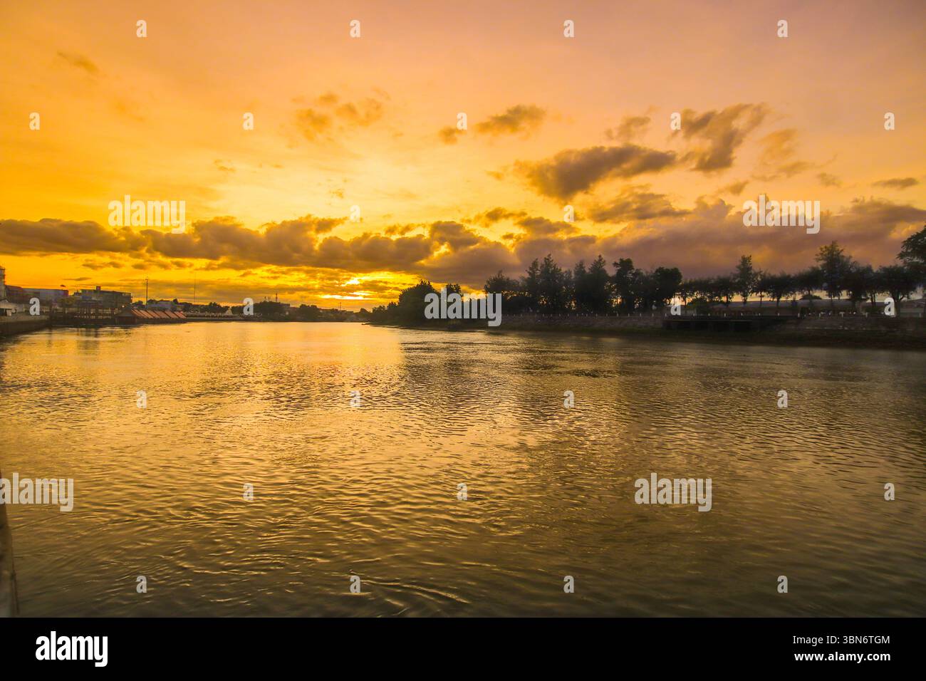 Ampia vista d'ora d'oro sul fiume con acqua illuminata dal sole, linea di alberi e morbide nuvole al crepuscolo, evocando calma e bellezza. Foto Stock