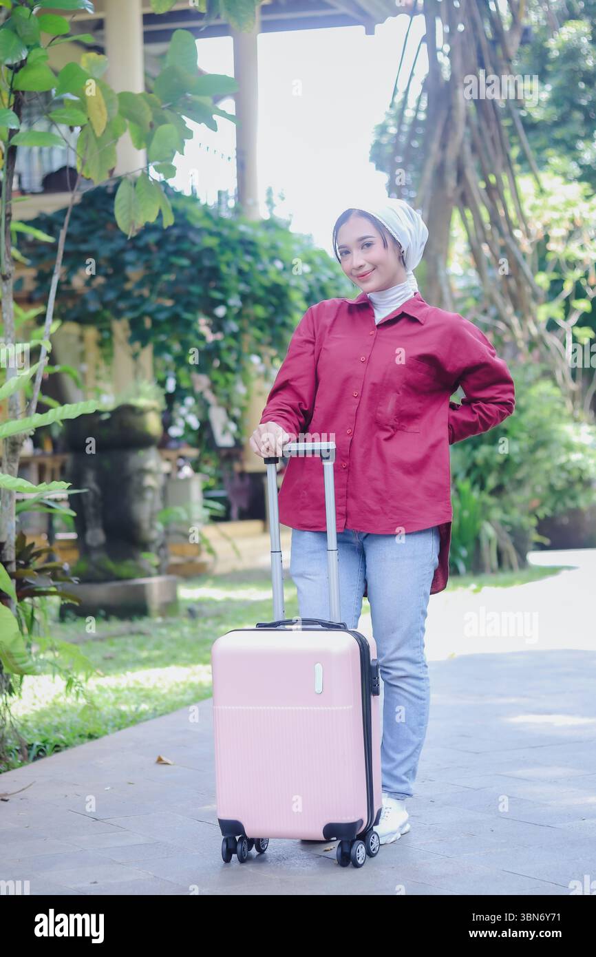 Una bella donna musulmana, indossando un elegante hijab, tira allegramente una valigia rosa, pronta per un viaggio emozionante in un ambiente delizioso, mostrando Foto Stock