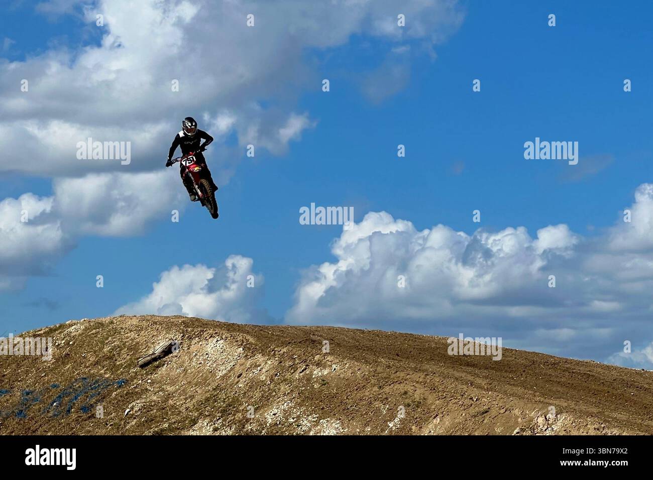 Pilota di Motocross che vola in alto su un enorme salto su una pista sterrata con un cielo azzurro e nuvole soffici Foto Stock