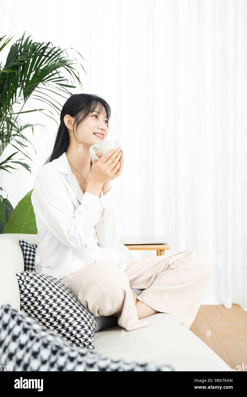Una giovane donna seduta seduta a gambe incrociate sul divano, tenendo una tazza di caffè con entrambe le mani Foto Stock