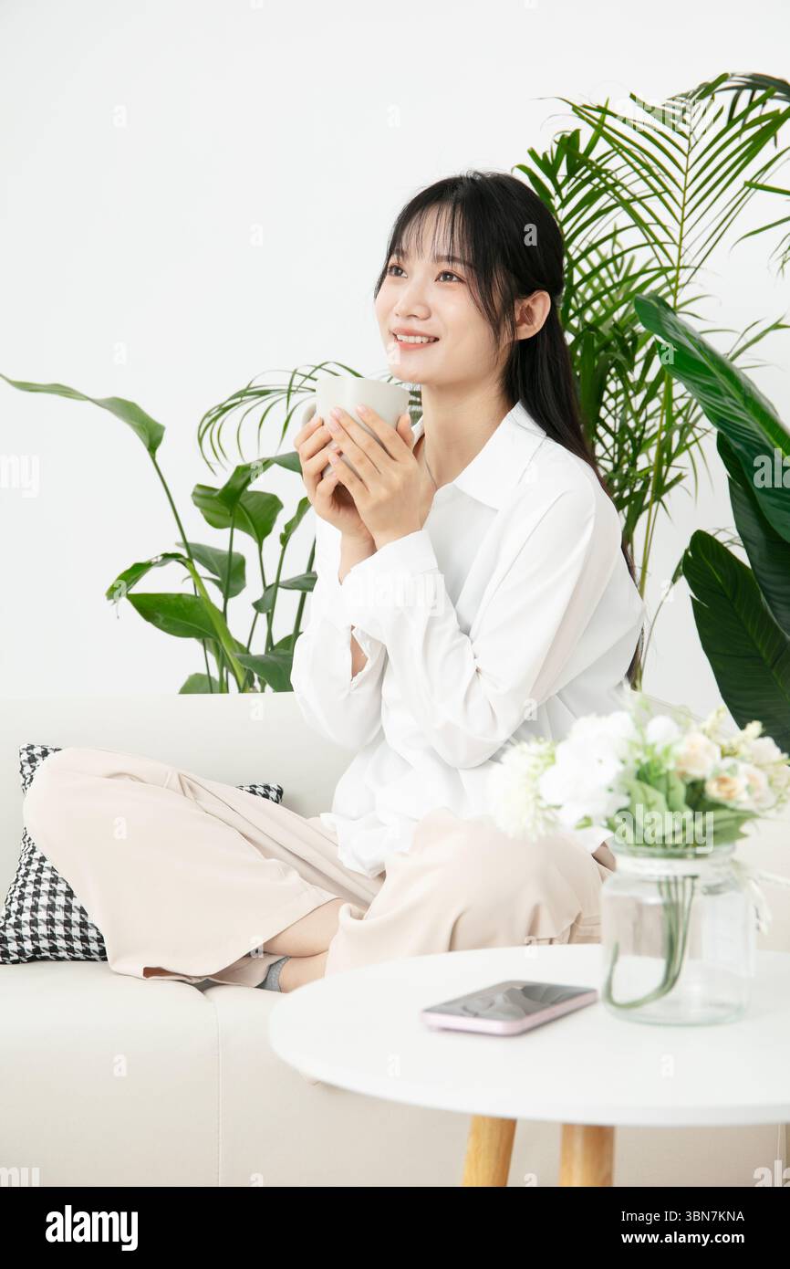 Una giovane donna seduta seduta a gambe incrociate sul divano, tenendo una tazza di caffè con entrambe le mani Foto Stock
