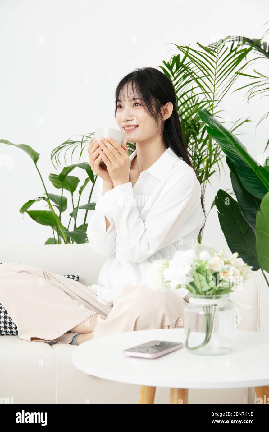 Una giovane donna seduta seduta a gambe incrociate sul divano, tenendo una tazza di caffè con entrambe le mani Foto Stock