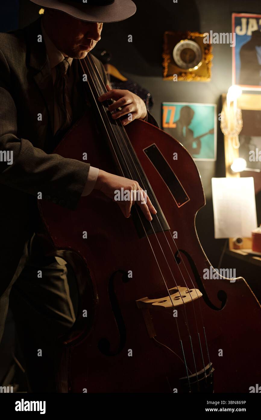 Uomo caucasico di mezza età che suona il basso verticale in un jazz club poco illuminato, indossa abiti e cappello, concentrandosi sullo strumento con poster jazz vintage e dischi in sottofondo Foto Stock