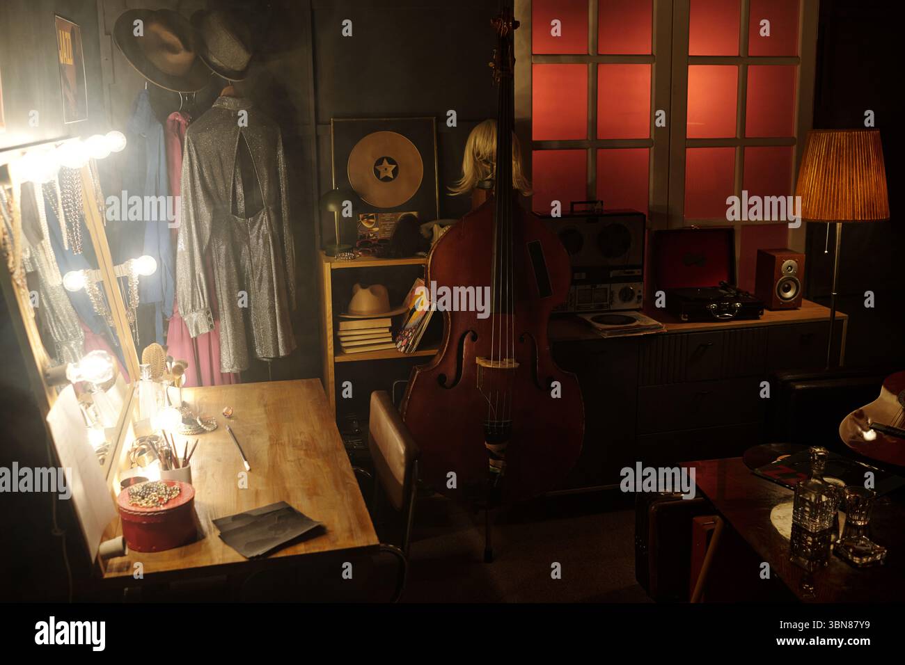 Spogliatoio per musicisti jazz vintage con contrabbasso in piedi vicino a un tavolo illuminato, un registratore retrò, dischi in vinile, cappelli e costumi da palco appesi sullo sfondo Foto Stock