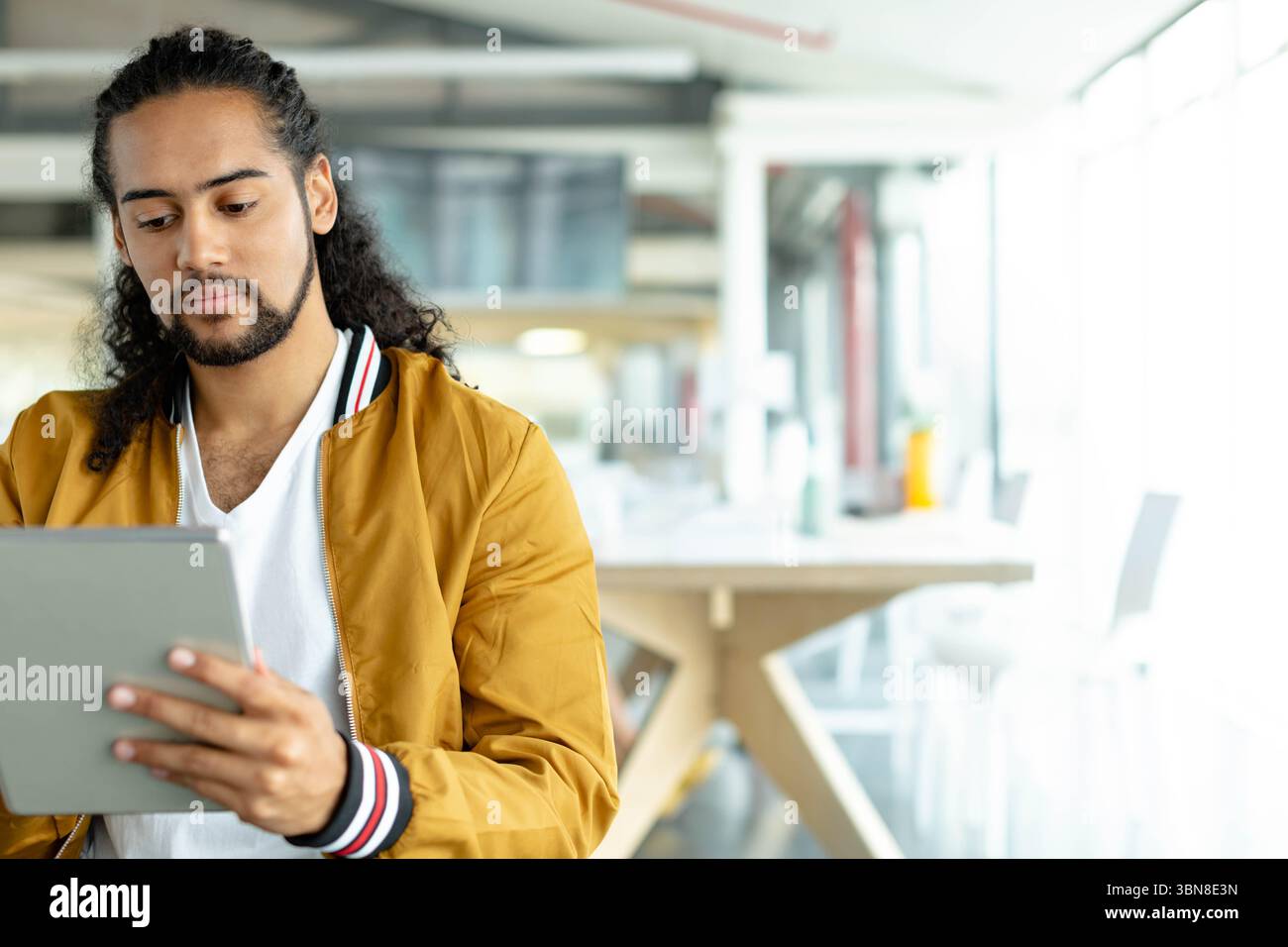 Uomo seduto nello spazio di coworking con un bomber che tiene un tablet vicino a finestre luminose, spazio per le copie Foto Stock
