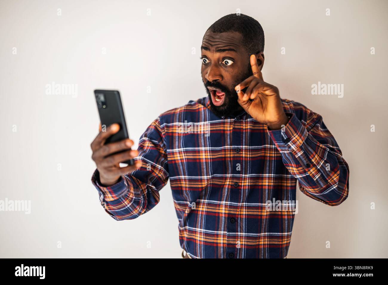 Uomo nero sorpreso e scioccato con una camicia accogliente davanti al muro e videochiamata sullo smartphone. Foto Stock
