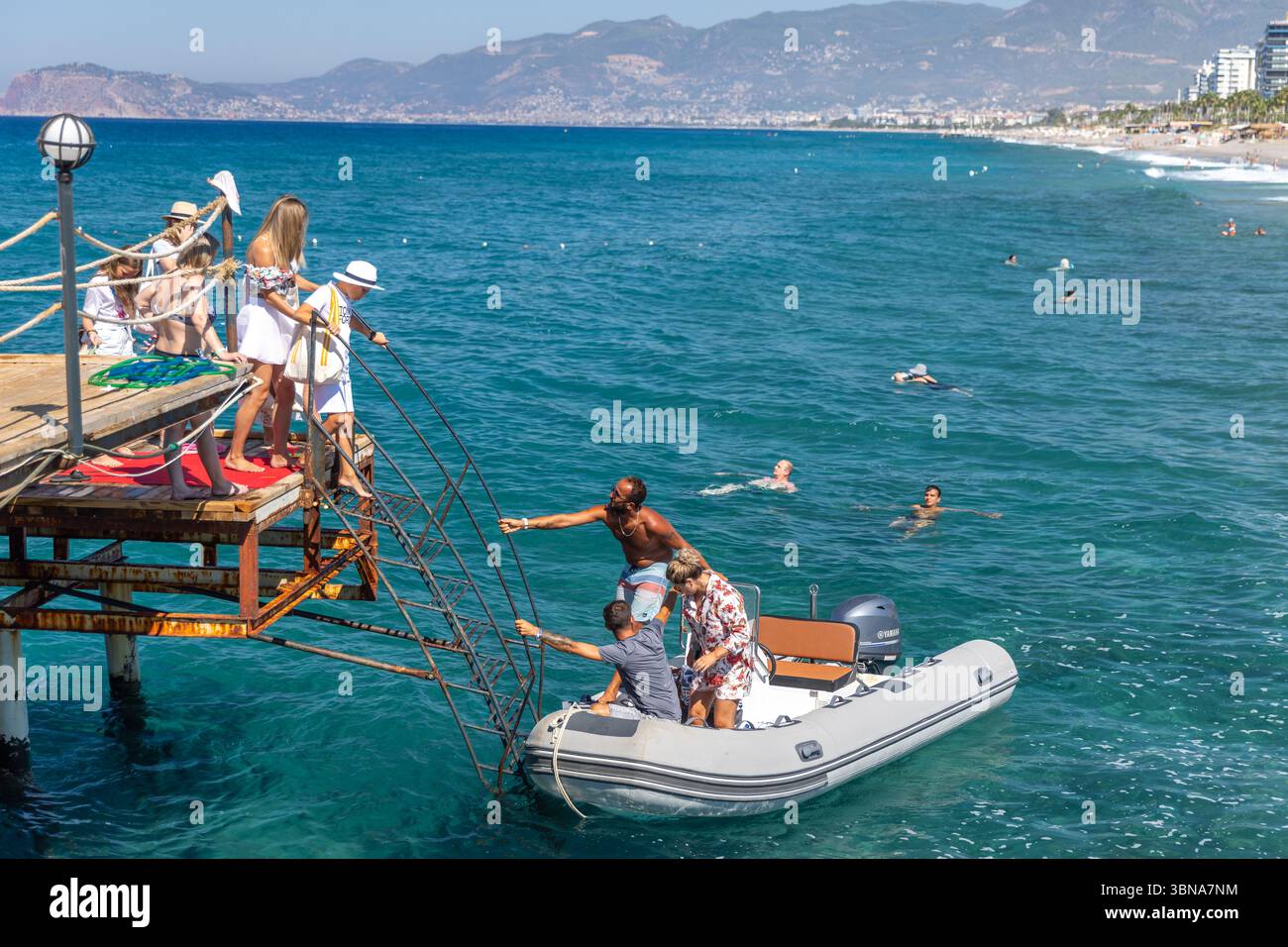 Mahmutlar, Turchia - 13 agosto 2024: Gruppo di persone che si trasferiscono da un molo panoramico a una barca a motore, godendosi il paesaggio costiero soleggiato e Foto Stock