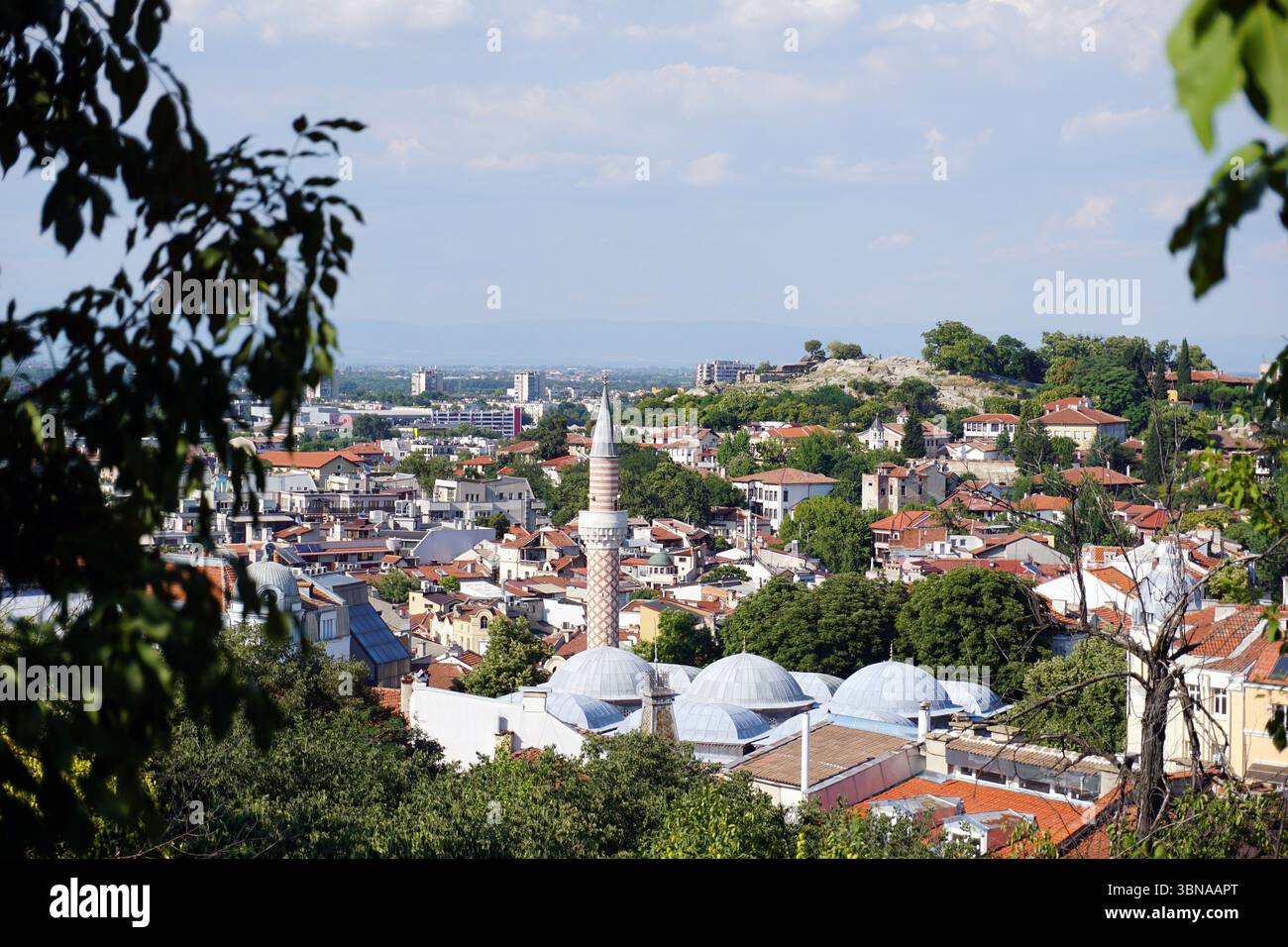 Plovdiv, la seconda città più grande della Bulgaria, è una delle città più antiche d’Europa continuamente abitate, con una storia che abbraccia oltre 8.000 anni. Incastonato lungo il fiume Maritsa, Un paesaggio urbano con una moschea con un alto minareto e diverse cupole, situato al centro della cornice. Il paesaggio urbano è un mix di edifici con tetti rossi e arancioni, intervallati da alberi e vegetazione. La città si estende in lontananza, con una collina o un'area sopraelevata visibile sullo sfondo. Il cielo è azzurro con alcune nuvole sparse. L'immagine viene scattata da un alto punto panoramico che offre una vista panoramica Foto Stock