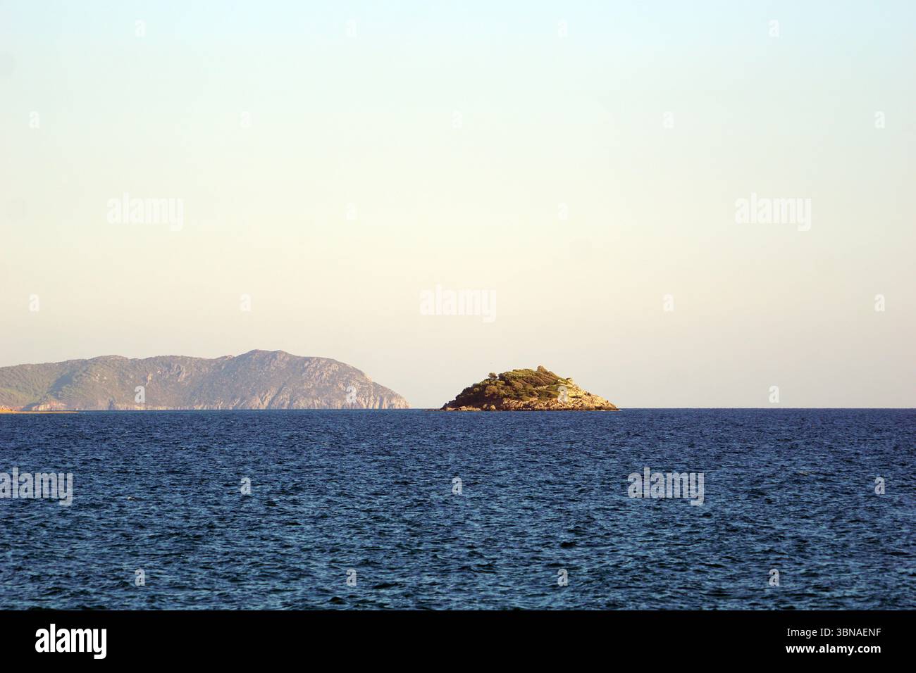 Una tranquilla scena di una piccola isola circondata da un vasto e profondo oceano blu, vicino a Bozyazi, Turchia. L'isola è coperta da una lussureggiante vegetazione verde e ha una costa rocciosa. In lontananza, una grande montagna rocciosa sorge dall'orizzonte, con la sua superficie aspra in contrasto con il mare calmo. Il cielo sopra è un azzurro chiaro e pallido, privo di nuvole. L'immagine viene scattata da lontano, fornendo una vista completa dell'isola e dell'ambiente circostante. L'occhio di un artista e la sua immaginazione Foto Stock