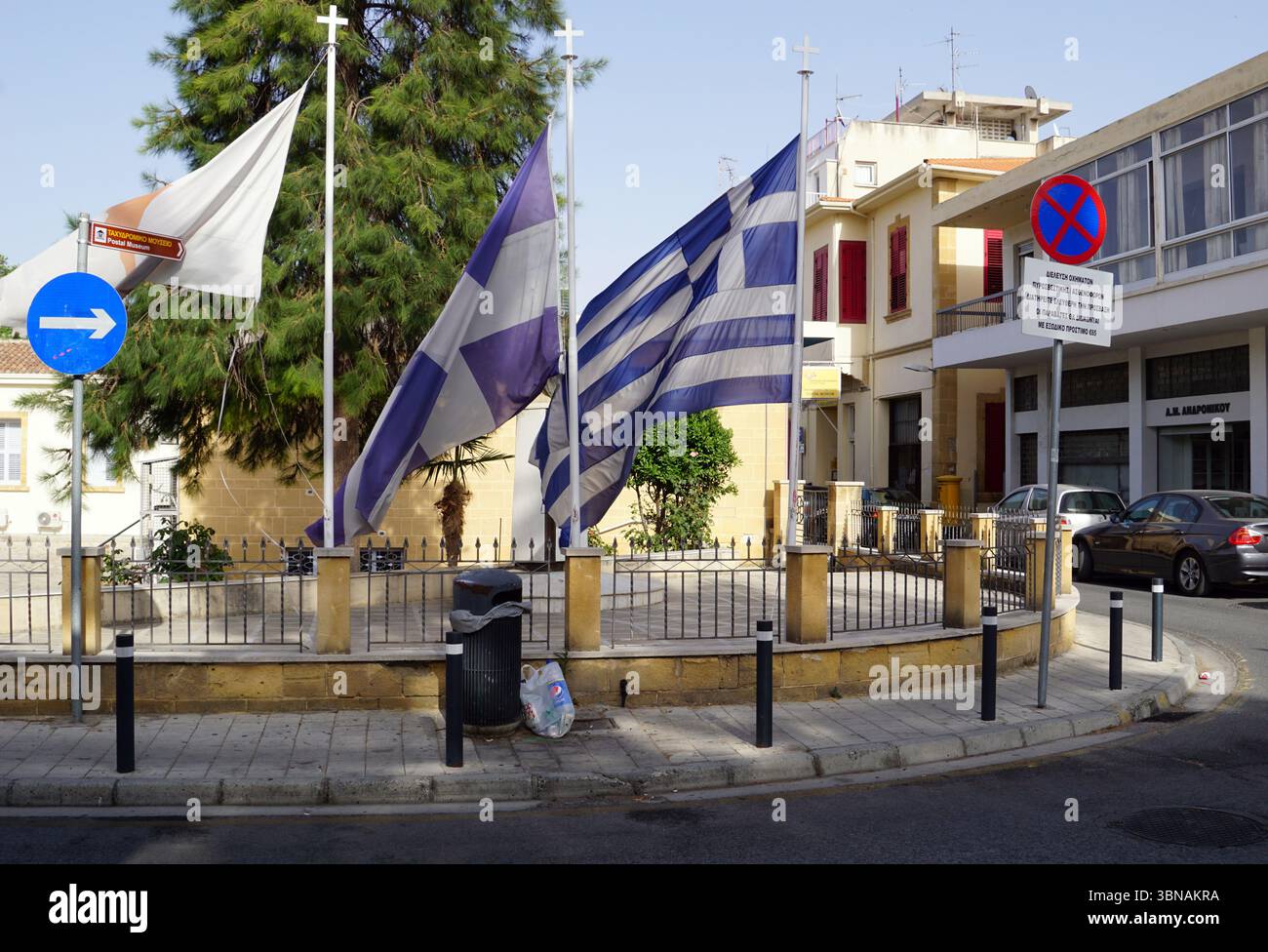 Nicosia (Lefkosia), Cipro, l'ultima capitale divisa del mondo, Un angolo di strada, dove tre bandiere sono ben visibili sui palchi. Una bandiera, con i suoi distintivi colori blu, bianco e viola, è posizionata a sinistra, mentre una bandiera bianca e blu è a destra. A sinistra è visibile anche una bandiera bianca e blu con una freccia bianca. Le bandiere sventolano nella brezza, aggiungendo un senso di movimento alla scena. Le bandiere sono fissate a paletti situati su un basso muro di pietra con una recinzione metallica nera. Vicino al muro si trova un cestino nero. Sullo sfondo, un edificio Foto Stock